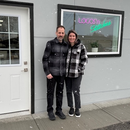 A man and woman stand proudly in front of their business, Logos N Stitches, showcasing their entrepreneurial spirit